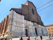 In der Julihitze empfängt Bologna die Reisenden mit engen Gassen, endlosen Arkaden und dem Duft von Kaffee aus jeder Bar. Und dann, auf der Piazza Maggiore, erhebt sich plötzlich ein riesiges Bauwerk – die Basilika San Petronio, die größte Kirche Bolognas und ein wahrer Riese, der bis heute unvollendet geblieben ist. Die Fassade erzählt eine eigene Geschichte Ihre Fassade erzählt eine eigene Geschichte: Der untere Teil glänzt mit weißem Marmor und Mustern, während der obere roh aus Ziegeln besteht. Sie wirkt dadurch ein wenig unvollständig – doch genau das macht ihren besonderen Charme aus. Es ist, als würde man ein Stück lebendige Geschichte sehen, halb vollendet, halb Traum. Der Bau begann bereits 1390. Die Bologneser wollten eine Kirche errichten, die sogar den Petersdom in Rom übertreffen sollte. Fast hätten sie es geschafft – San Petronio gehört zu den zehn größten Kirchen der Welt. Doch der Papst bemerkte schnell den Ehrgeiz der Stadt und stoppte die gewaltigen Pläne: „Rom ist das Zentrum, Bologna soll zweitrangig bleiben.“ So blieb sie: großartig, aber unvollendet. Kleiderordnung in der Basilika muss beachtet werden Im Inneren empfängt einen kühle Weite. Hohe Gewölbe, bunte Glasfenster, das Licht, das von oben fällt – und das Gefühl, dass hier immer Platz für Stille bleibt. In der Kirche gibt es viele Kunstschätze, doch am meisten erstaunt die Meridianlinie – ein schmaler Messingstreifen, der in den Boden eingelassen ist. Durch ein kleines Loch in der Decke markiert die Sonne täglich die Zeit und die Jahreszeit. Im 17. Jahrhundert war dies eine Art astronomisches Observatorium! Wir traten ein, während draußen die Hitze drückte. Am Eingang kontrollieren Helfer die Kleidung: Hier gelten, wie in den meisten großen Kirchen Italiens, Regeln der Anständigkeit. Wer Shorts oder Röcke trägt, die zu kurz sind, bekommt leichte Schals zum Bedecken. Sie sind durchsichtig, aber ausreichend – und so sehen plötzlich alle Touristen ein wenig gleich aus, als gehörten sie zu einem Klub von Reisenden auf der Suche nach Kühle und Schönheit. Pause auf den Treppen der Basilika Drinnen erlebten wir eine Messe auf Polnisch. Die Stimmen des Chors hallten unter den gotischen Gewölben wider, und es war eigenartig und schön, eine vertraute slawische Sprache im Herzen einer italienischen Stadt zu hören. Der schönste Moment kam, als wir hinausgingen. Die Hitze schlug uns ins Gesicht, und wie viele andere setzten wir uns auf die breiten Stufen vor der Basilika. Hier ruht immer jemand: Touristen mit Rucksäcken, Einheimische mit Eis, Studenten mit Gitarren. Ein wenig Schatten – und man spürt, dass man im Rhythmus der Stadt mitschwingt. Geschichte und Gegenwart fließen zusammen San Petronio ist nicht nur ein architektonisches Denkmal. Sie ist ein Symbol dafür, wie Bologna immer nach mehr strebte, als man ihr zugestand. Ein Ort, an dem Geschichte und Gegenwart zusammenfließen: der alte Traum von Größe, Gebete in vielen Sprachen, Lieder auf dem Platz und der lebendige Puls der Stadt. Anreise zur Basilika San Petronio 📍 Adresse: Piazza Maggiore, Bologna, Italien. 🚆 Vom Bahnhof Bologna Centrale: Zu Fuß sind es etwa 20 Minuten. Am einfachsten geht man die Via dell’Indipendenza entlang – die Hauptstraße, die direkt ins Zentrum führt. 🚌 Wer nicht laufen möchte, kann auch den Bus nehmen (z. B. Nr. 25, 27 oder 30). Aussteigen sollte man an der Haltestelle Rizzoli, die nur wenige Schritte vom Platz entfernt liegt. 🚶‍♀️ Befindet man sich bereits im Zentrum von Bologna, kann man sie unmöglich übersehen: Die Basilika beherrscht die gesamte Piazza.