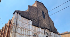 In der Julihitze empfängt Bologna die Reisenden mit engen Gassen, endlosen Arkaden und dem Duft von Kaffee aus jeder Bar. Und dann, auf der Piazza Maggiore, erhebt sich plötzlich ein riesiges Bauwerk – die Basilika San Petronio, die größte Kirche Bolognas und ein wahrer Riese, der bis heute unvollendet geblieben ist. Die Fassade erzählt eine eigene Geschichte Ihre Fassade erzählt eine eigene Geschichte: Der untere Teil glänzt mit weißem Marmor und Mustern, während der obere roh aus Ziegeln besteht. Sie wirkt dadurch ein wenig unvollständig – doch genau das macht ihren besonderen Charme aus. Es ist, als würde man ein Stück lebendige Geschichte sehen, halb vollendet, halb Traum. Der Bau begann bereits 1390. Die Bologneser wollten eine Kirche errichten, die sogar den Petersdom in Rom übertreffen sollte. Fast hätten sie es geschafft – San Petronio gehört zu den zehn größten Kirchen der Welt. Doch der Papst bemerkte schnell den Ehrgeiz der Stadt und stoppte die gewaltigen Pläne: „Rom ist das Zentrum, Bologna soll zweitrangig bleiben.“ So blieb sie: großartig, aber unvollendet. Kleiderordnung in der Basilika muss beachtet werden Im Inneren empfängt einen kühle Weite. Hohe Gewölbe, bunte Glasfenster, das Licht, das von oben fällt – und das Gefühl, dass hier immer Platz für Stille bleibt. In der Kirche gibt es viele Kunstschätze, doch am meisten erstaunt die Meridianlinie – ein schmaler Messingstreifen, der in den Boden eingelassen ist. Durch ein kleines Loch in der Decke markiert die Sonne täglich die Zeit und die Jahreszeit. Im 17. Jahrhundert war dies eine Art astronomisches Observatorium! Wir traten ein, während draußen die Hitze drückte. Am Eingang kontrollieren Helfer die Kleidung: Hier gelten, wie in den meisten großen Kirchen Italiens, Regeln der Anständigkeit. Wer Shorts oder Röcke trägt, die zu kurz sind, bekommt leichte Schals zum Bedecken. Sie sind durchsichtig, aber ausreichend – und so sehen plötzlich alle Touristen ein wenig gleich aus, als gehörten sie zu einem Klub von Reisenden auf der Suche nach Kühle und Schönheit. Pause auf den Treppen der Basilika Drinnen erlebten wir eine Messe auf Polnisch. Die Stimmen des Chors hallten unter den gotischen Gewölben wider, und es war eigenartig und schön, eine vertraute slawische Sprache im Herzen einer italienischen Stadt zu hören. Der schönste Moment kam, als wir hinausgingen. Die Hitze schlug uns ins Gesicht, und wie viele andere setzten wir uns auf die breiten Stufen vor der Basilika. Hier ruht immer jemand: Touristen mit Rucksäcken, Einheimische mit Eis, Studenten mit Gitarren. Ein wenig Schatten – und man spürt, dass man im Rhythmus der Stadt mitschwingt. Geschichte und Gegenwart fließen zusammen San Petronio ist nicht nur ein architektonisches Denkmal. Sie ist ein Symbol dafür, wie Bologna immer nach mehr strebte, als man ihr zugestand. Ein Ort, an dem Geschichte und Gegenwart zusammenfließen: der alte Traum von Größe, Gebete in vielen Sprachen, Lieder auf dem Platz und der lebendige Puls der Stadt. Anreise zur Basilika San Petronio 📍 Adresse: Piazza Maggiore, Bologna, Italien. 🚆 Vom Bahnhof Bologna Centrale: Zu Fuß sind es etwa 20 Minuten. Am einfachsten geht man die Via dell’Indipendenza entlang – die Hauptstraße, die direkt ins Zentrum führt. 🚌 Wer nicht laufen möchte, kann auch den Bus nehmen (z. B. Nr. 25, 27 oder 30). Aussteigen sollte man an der Haltestelle Rizzoli, die nur wenige Schritte vom Platz entfernt liegt. 🚶‍♀️ Befindet man sich bereits im Zentrum von Bologna, kann man sie unmöglich übersehen: Die Basilika beherrscht die gesamte Piazza.