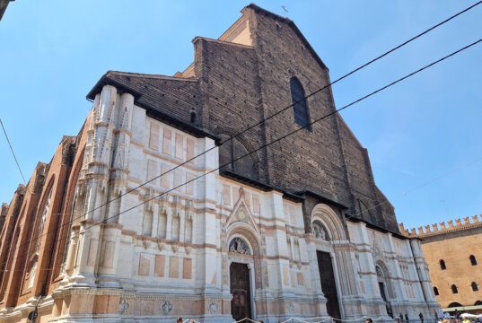In der Julihitze empfängt Bologna die Reisenden mit engen Gassen, endlosen Arkaden und dem Duft von Kaffee aus jeder Bar. Und dann, auf der Piazza Maggiore, erhebt sich plötzlich ein riesiges Bauwerk – die Basilika San Petronio, die größte Kirche Bolognas und ein wahrer Riese, der bis heute unvollendet geblieben ist. Die Fassade erzählt eine eigene Geschichte Ihre Fassade erzählt eine eigene Geschichte: Der untere Teil glänzt mit weißem Marmor und Mustern, während der obere roh aus Ziegeln besteht. Sie wirkt dadurch ein wenig unvollständig – doch genau das macht ihren besonderen Charme aus. Es ist, als würde man ein Stück lebendige Geschichte sehen, halb vollendet, halb Traum. Der Bau begann bereits 1390. Die Bologneser wollten eine Kirche errichten, die sogar den Petersdom in Rom übertreffen sollte. Fast hätten sie es geschafft – San Petronio gehört zu den zehn größten Kirchen der Welt. Doch der Papst bemerkte schnell den Ehrgeiz der Stadt und stoppte die gewaltigen Pläne: „Rom ist das Zentrum, Bologna soll zweitrangig bleiben.“ So blieb sie: großartig, aber unvollendet. Kleiderordnung in der Basilika muss beachtet werden Im Inneren empfängt einen kühle Weite. Hohe Gewölbe, bunte Glasfenster, das Licht, das von oben fällt – und das Gefühl, dass hier immer Platz für Stille bleibt. In der Kirche gibt es viele Kunstschätze, doch am meisten erstaunt die Meridianlinie – ein schmaler Messingstreifen, der in den Boden eingelassen ist. Durch ein kleines Loch in der Decke markiert die Sonne täglich die Zeit und die Jahreszeit. Im 17. Jahrhundert war dies eine Art astronomisches Observatorium! Wir traten ein, während draußen die Hitze drückte. Am Eingang kontrollieren Helfer die Kleidung: Hier gelten, wie in den meisten großen Kirchen Italiens, Regeln der Anständigkeit. Wer Shorts oder Röcke trägt, die zu kurz sind, bekommt leichte Schals zum Bedecken. Sie sind durchsichtig, aber ausreichend – und so sehen plötzlich alle Touristen ein wenig gleich aus, als gehörten sie zu einem Klub von Reisenden auf der Suche nach Kühle und Schönheit. Pause auf den Treppen der Basilika Drinnen erlebten wir eine Messe auf Polnisch. Die Stimmen des Chors hallten unter den gotischen Gewölben wider, und es war eigenartig und schön, eine vertraute slawische Sprache im Herzen einer italienischen Stadt zu hören. Der schönste Moment kam, als wir hinausgingen. Die Hitze schlug uns ins Gesicht, und wie viele andere setzten wir uns auf die breiten Stufen vor der Basilika. Hier ruht immer jemand: Touristen mit Rucksäcken, Einheimische mit Eis, Studenten mit Gitarren. Ein wenig Schatten – und man spürt, dass man im Rhythmus der Stadt mitschwingt. Geschichte und Gegenwart fließen zusammen San Petronio ist nicht nur ein architektonisches Denkmal. Sie ist ein Symbol dafür, wie Bologna immer nach mehr strebte, als man ihr zugestand. Ein Ort, an dem Geschichte und Gegenwart zusammenfließen: der alte Traum von Größe, Gebete in vielen Sprachen, Lieder auf dem Platz und der lebendige Puls der Stadt. Anreise zur Basilika San Petronio 📍 Adresse: Piazza Maggiore, Bologna, Italien. 🚆 Vom Bahnhof Bologna Centrale: Zu Fuß sind es etwa 20 Minuten. Am einfachsten geht man die Via dell’Indipendenza entlang – die Hauptstraße, die direkt ins Zentrum führt. 🚌 Wer nicht laufen möchte, kann auch den Bus nehmen (z. B. Nr. 25, 27 oder 30). Aussteigen sollte man an der Haltestelle Rizzoli, die nur wenige Schritte vom Platz entfernt liegt. 🚶‍♀️ Befindet man sich bereits im Zentrum von Bologna, kann man sie unmöglich übersehen: Die Basilika beherrscht die gesamte Piazza.