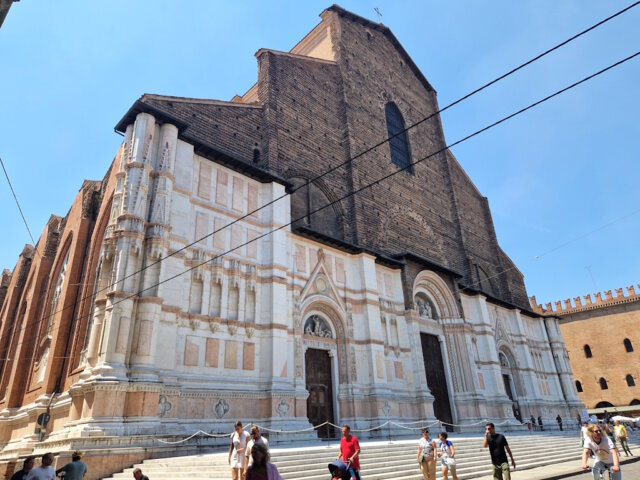 basilika-san-petronio-bologna (1) In der Julihitze empfängt Bologna die Reisenden mit engen Gassen, endlosen Arkaden und dem Duft von Kaffee aus jeder Bar. Und dann, auf der Piazza Maggiore, erhebt sich plötzlich ein riesiges Bauwerk – die Basilika San Petronio, die größte Kirche Bolognas und ein wahrer Riese, der bis heute unvollendet geblieben ist. Die Fassade erzählt eine eigene Geschichte Ihre Fassade erzählt eine eigene Geschichte: Der untere Teil glänzt mit weißem Marmor und Mustern, während der obere roh aus Ziegeln besteht. Sie wirkt dadurch ein wenig unvollständig – doch genau das macht ihren besonderen Charme aus. Es ist, als würde man ein Stück lebendige Geschichte sehen, halb vollendet, halb Traum. Der Bau begann bereits 1390. Die Bologneser wollten eine Kirche errichten, die sogar den Petersdom in Rom übertreffen sollte. Fast hätten sie es geschafft – San Petronio gehört zu den zehn größten Kirchen der Welt. Doch der Papst bemerkte schnell den Ehrgeiz der Stadt und stoppte die gewaltigen Pläne: „Rom ist das Zentrum, Bologna soll zweitrangig bleiben.“ So blieb sie: großartig, aber unvollendet. Kleiderordnung in der Basilika muss beachtet werden Im Inneren empfängt einen kühle Weite. Hohe Gewölbe, bunte Glasfenster, das Licht, das von oben fällt – und das Gefühl, dass hier immer Platz für Stille bleibt. In der Kirche gibt es viele Kunstschätze, doch am meisten erstaunt die Meridianlinie – ein schmaler Messingstreifen, der in den Boden eingelassen ist. Durch ein kleines Loch in der Decke markiert die Sonne täglich die Zeit und die Jahreszeit. Im 17. Jahrhundert war dies eine Art astronomisches Observatorium! Wir traten ein, während draußen die Hitze drückte. Am Eingang kontrollieren Helfer die Kleidung: Hier gelten, wie in den meisten großen Kirchen Italiens, Regeln der Anständigkeit. Wer Shorts oder Röcke trägt, die zu kurz sind, bekommt leichte Schals zum Bedecken. Sie sind durchsichtig, aber ausreichend – und so sehen plötzlich alle Touristen ein wenig gleich aus, als gehörten sie zu einem Klub von Reisenden auf der Suche nach Kühle und Schönheit. Pause auf den Treppen der Basilika Drinnen erlebten wir eine Messe auf Polnisch. Die Stimmen des Chors hallten unter den gotischen Gewölben wider, und es war eigenartig und schön, eine vertraute slawische Sprache im Herzen einer italienischen Stadt zu hören. Der schönste Moment kam, als wir hinausgingen. Die Hitze schlug uns ins Gesicht, und wie viele andere setzten wir uns auf die breiten Stufen vor der Basilika. Hier ruht immer jemand: Touristen mit Rucksäcken, Einheimische mit Eis, Studenten mit Gitarren. Ein wenig Schatten – und man spürt, dass man im Rhythmus der Stadt mitschwingt. Geschichte und Gegenwart fließen zusammen San Petronio ist nicht nur ein architektonisches Denkmal. Sie ist ein Symbol dafür, wie Bologna immer nach mehr strebte, als man ihr zugestand. Ein Ort, an dem Geschichte und Gegenwart zusammenfließen: der alte Traum von Größe, Gebete in vielen Sprachen, Lieder auf dem Platz und der lebendige Puls der Stadt. Anreise zur Basilika San Petronio 📍 Adresse: Piazza Maggiore, Bologna, Italien. 🚆 Vom Bahnhof Bologna Centrale: Zu Fuß sind es etwa 20 Minuten. Am einfachsten geht man die Via dell’Indipendenza entlang – die Hauptstraße, die direkt ins Zentrum führt. 🚌 Wer nicht laufen möchte, kann auch den Bus nehmen (z. B. Nr. 25, 27 oder 30). Aussteigen sollte man an der Haltestelle Rizzoli, die nur wenige Schritte vom Platz entfernt liegt. 🚶♀️ Befindet man sich bereits im Zentrum von Bologna, kann man sie unmöglich übersehen: Die Basilika beherrscht die gesamte Piazza.