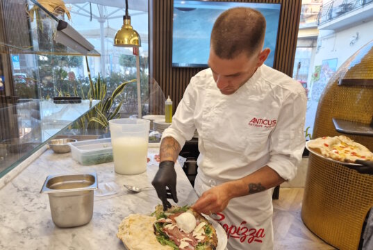 Finaler Handgriff an der Nuda e Cruda: Der vorgebackene Pizzateig wird im Anticus mit frischen Zutaten wie Büffelmozzarella, Rucola und Prosciutto belegt – ein kreatives Gericht zwischen Pizza und gefüllter Focaccia. (Foto: © Bastian Glumm)
